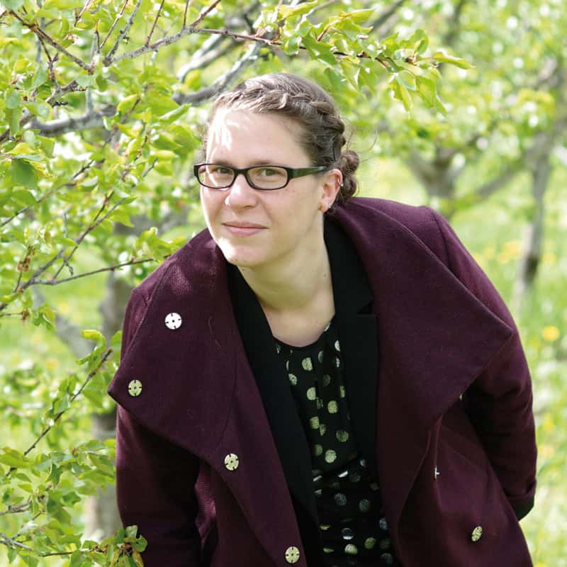 Porträtfoto Lara Schink, draußen im grünen Park, Lara steht neben einem grünen Strauch und schaut direkt in die Kamera