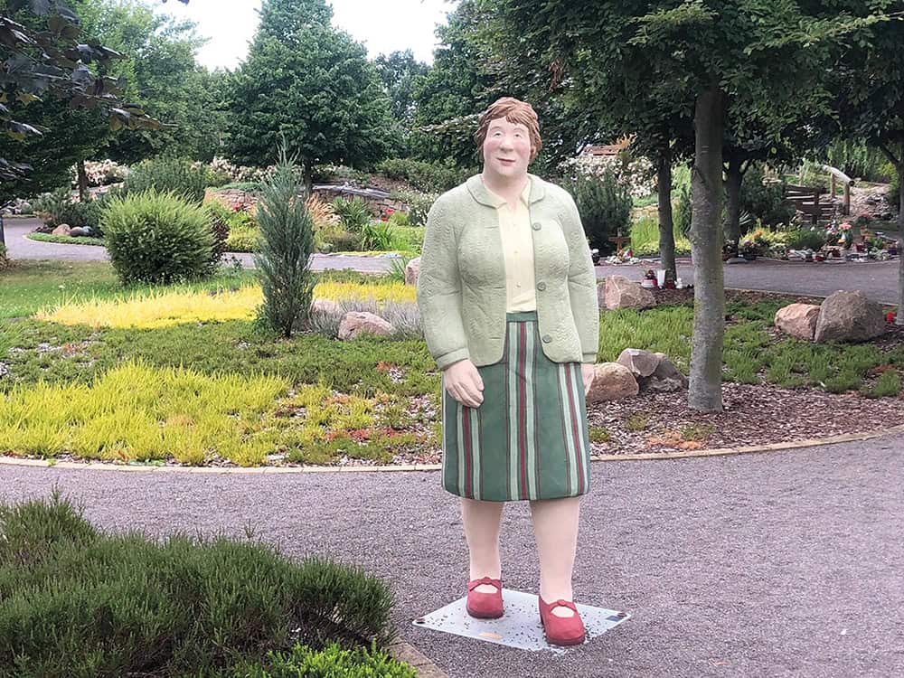 Alltagsmensch im Friedgarten, ältere Frau steht in der Mitte auf dem Kiesweg und schaut und die Kamera