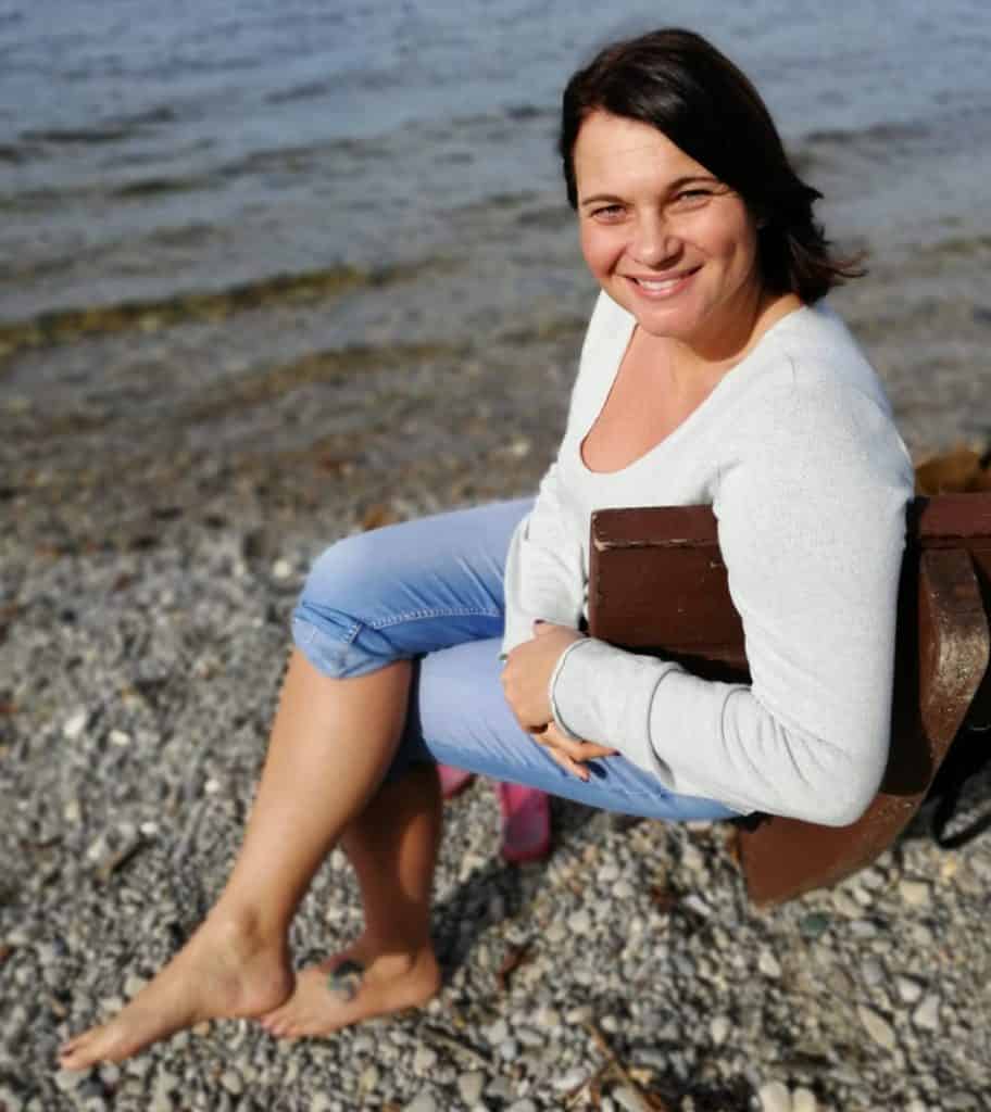 Porträtfoto Katrin Gärtner, Katrin sitzt auf einer Bank am Strand mit Kies und lächelt in die Kamera