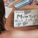 Kind/Mädchen sitzt an einem Tisch und malt. Man sieht ihr über die rechte Schulter und sieht ihr gemaltes in einem Block: „Mein Papi ist tot” steht in grau geschrieben mit grauen Herzen darum.