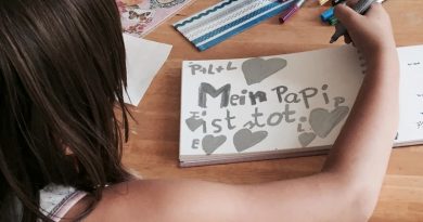 Kind/Mädchen sitzt an einem Tisch und malt. Man sieht ihr über die rechte Schulter und sieht ihr gemaltes in einem Block: „Mein Papi ist tot” steht in grau geschrieben mit grauen Herzen darum.