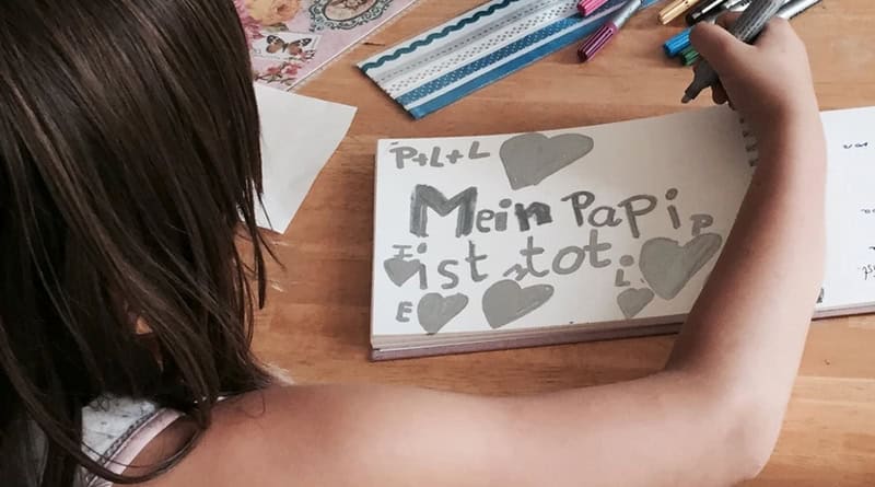 Kind/Mädchen sitzt an einem Tisch und malt. Man sieht ihr über die rechte Schulter und sieht ihr gemaltes in einem Block: „Mein Papi ist tot” steht in grau geschrieben mit grauen Herzen darum.