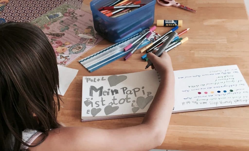 Kind/Mädchen sitzt an einem Tisch und malt. Man sieht ihr über die rechte Schulter und sieht ihr gemaltes in einem Block: „Mein Papi ist tot” steht in grau geschrieben mit grauen Herzen darum.