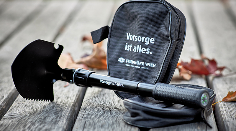 Foto schwarze Klappschaufel mit der Aufschrift „Vorsorge ist alles” als Merchandise, dahinter die Schaufel-Verpackung mit gleicher Aufschrift