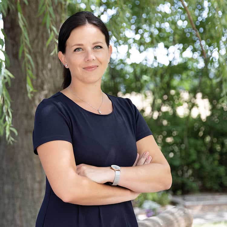 Porträtfoto Kristin Rogge, Kristin steht mit verschränkten Armen in der Natur vor einem Baum