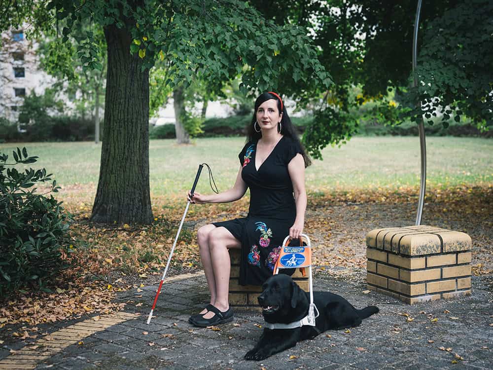 Jennifer Sonntag Porträtfoto. Jenny sitzt in einem Park unter einem Baum auf einem Stein. Ihr Hund Paul liegt in Platzhaltung vor ihr.