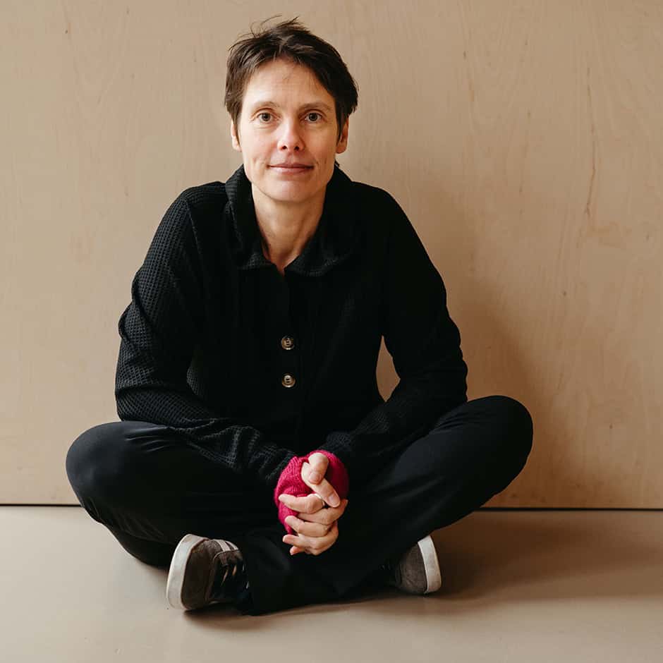 Porträtfoto Barbara Rolf von Foto: Isabell Steinert, Dazwischen, Freiburg, Barbara sitzt im Schneidersitz vor einer beigen Wand