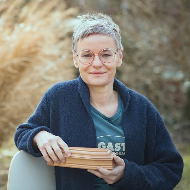 Portraitfoto Christine Pernlochner-Kügler von Gerhard Berger, Christine sitzt in der Natur, hält einen kleinen Holzsarg in der Hand und lächelt in die Kamera