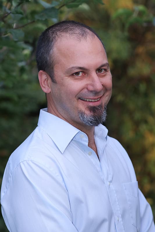 Portraitfoto Denis Bicanic, Denis lächelt seitlich in die Kamera, er trägt ein weiß-hellblaues Hemd