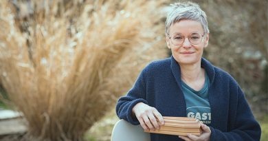 Portraitfoto Christine Pernlochner-Kügler von Gerhard Berger, Christine sitzt in der Natur, hält einen kleinen Holzsarg in der Hand und lächelt in die Kamera