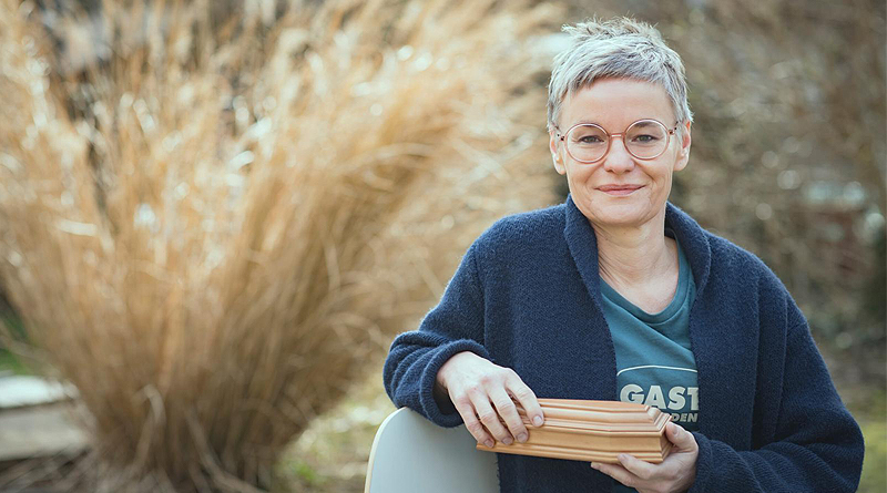 Portraitfoto Christine Pernlochner-Kügler von Gerhard Berger, Christine sitzt in der Natur, hält einen kleinen Holzsarg in der Hand und lächelt in die Kamera