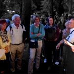 Menschen auf einem Friedhof bei einer Veranstaltung, sie stehen im Kreis und schauen auf die Frau mit Mikro, die etwas vorliest