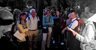 Menschen auf einem Friedhof bei einer Veranstaltung, sie stehen im Kreis und schauen auf die Frau mit Mikro, die etwas vorliest