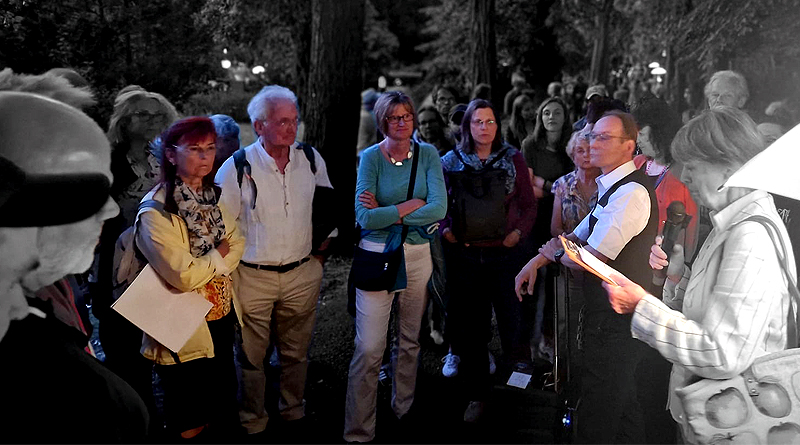 Menschen auf einem Friedhof bei einer Veranstaltung, sie stehen im Kreis und schauen auf die Frau mit Mikro, die etwas vorliest