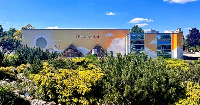 Flamarium, Außenfassade, davor Sträucher, ein Teich, blauer Himmel und Sonnenschein