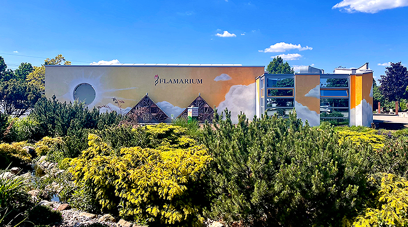 Flamarium, Außenfassade, davor Sträucher, ein Teich, blauer Himmel und Sonnenschein