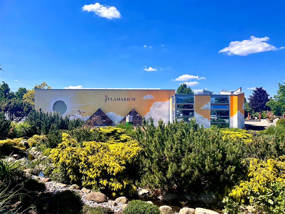 Flamarium, Außenfassade, davor Sträucher, ein Teich, blauer Himmel und Sonnenschein